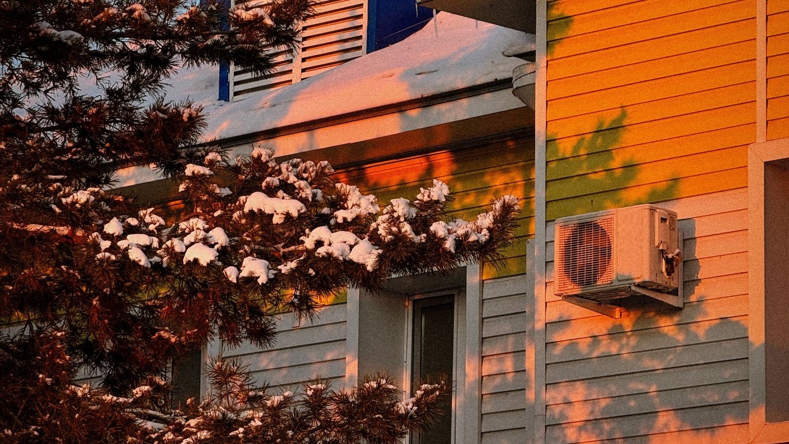 Wall-mounted ductless heat pump outdoor unit on a snow-covered house at sunset, a cold climate mini split running in winter
